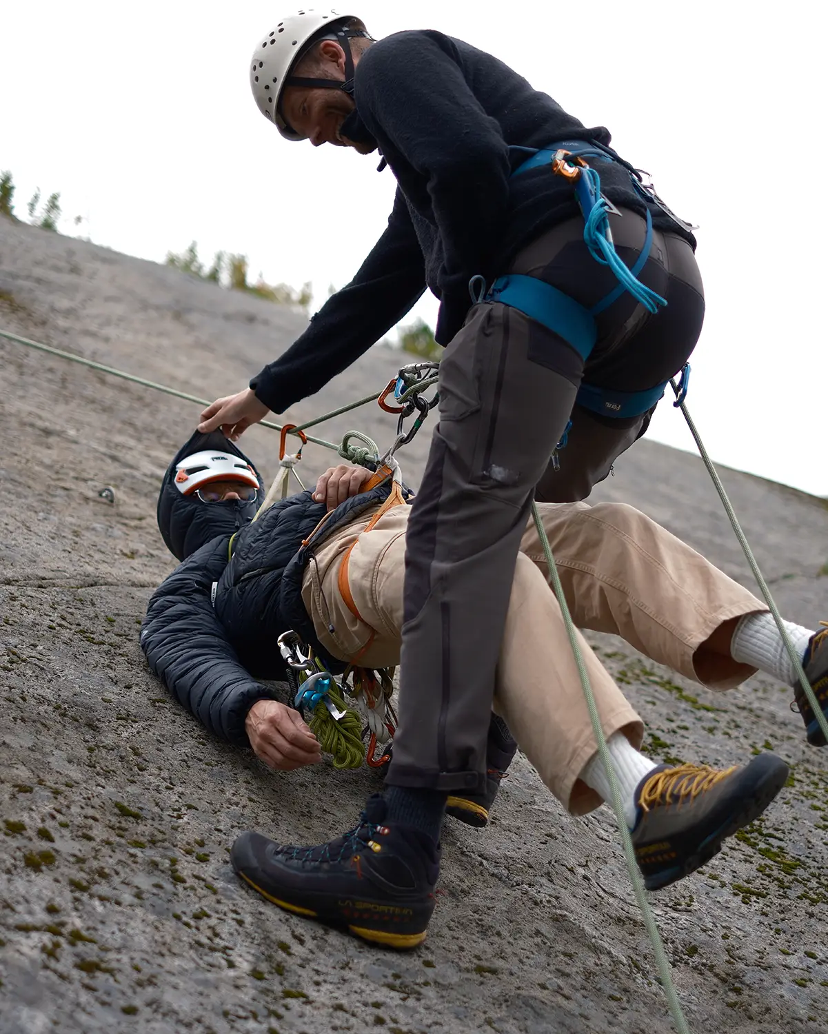 Climber lying on a rocky slope while a partner in helmet and harness assists with ropes for a rescue or belay.