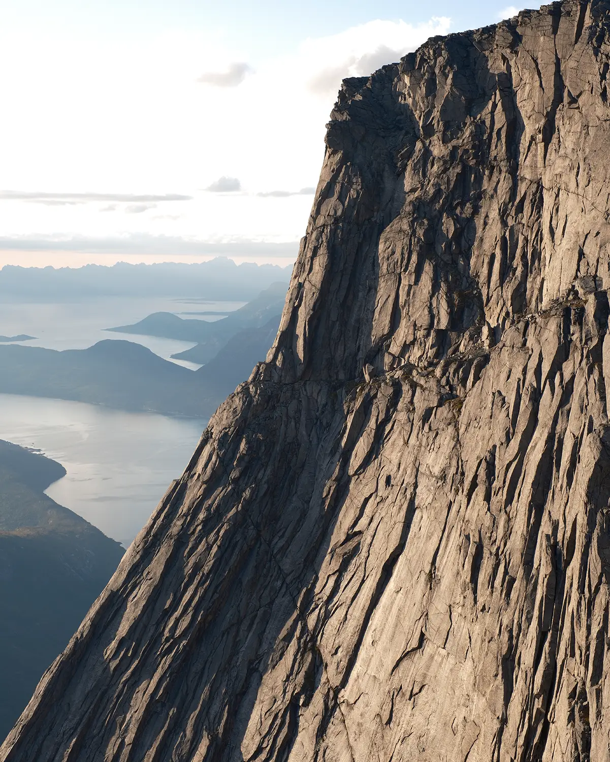 Steep rocky cliff dominates the right, with a fjord and distant mountains on the left under a clear sky and calm water.